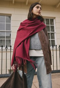 Brauner Mantel über einem grauen Pullover, kombiniert mit hellen Jeans. Ein lebhaft roter Schal fällt elegant mit fransigen Enden. Eine dunkle Tasche wird gehalten.