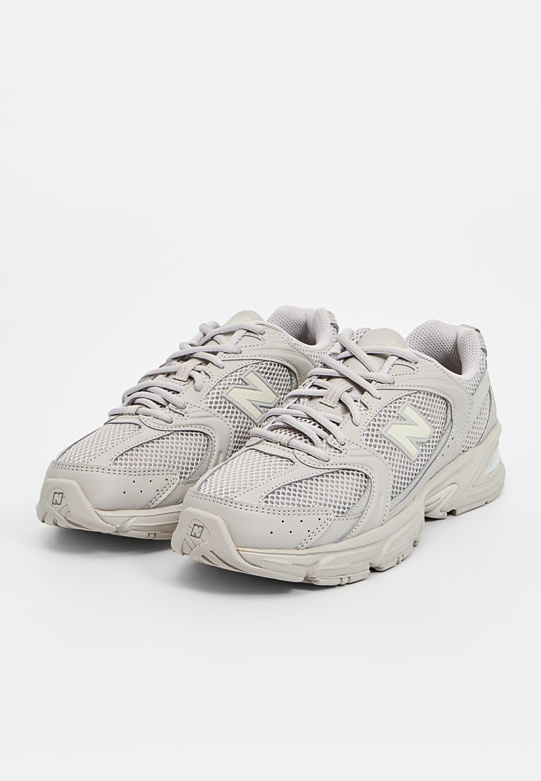 Zapatillas deportivas gris claro fabricadas con malla y materiales sintéticos. Presentan una superficie texturizada, diseño con cordones y una suela acolchada.