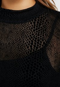 Transparent svart mesh topp med prickmönster, strukturerad tyg och hög krage. Utmärkande genomskinliga partier skapar visuell kontrast.