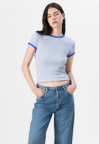 Modella con lunghi capelli castani, che indossa una camicia a maniche corte a righe e jeans blu a vita alta, in piedi davanti a uno sfondo bianco.