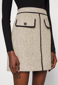 Jupe en tweed beige avec texture, bordure noire et deux poches avant ornées de boutons noirs. Coupe droite avec un ourlet légèrement évasé.