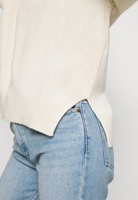 Cremfarbener Strickpullover mit geripptem Saum und seitlichen Schlitzen, kombiniert mit hellblauen, eng geschnittenen Jeans. Die Textur wirkt weich und atmungsaktiv.