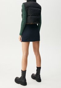 Czarna puchowa kamizelka, zielony prążkowany sweter, granatowa mini spódniczka w prążki oraz czarne botki za kostkę z grubymi podeszwami i zamkami, widziane od tyłu.