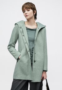 Lysegrønn ullblandingsfrakk med hette, med glidelås og trykknapp, to fremre lommer og teksturert stoff.