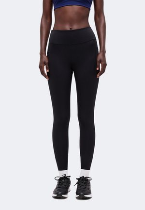 Kvinde iført sorte leggings med høj talje, hvide sokker og sorte sportssko, stående mod en ensfarvet lys baggrund.