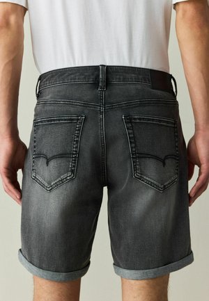 Back view of person wearing black washed denim shorts with rolled cuffs and a white tucked-in t-shirt.