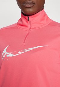 Ružičasta dugog rukava majica Nike s visokim ovratnikom. Sadrži bijelu Nike swoosh oznaku otisnutu na prednjem dijelu. Izrađena od glatkog, laganog materijala.
