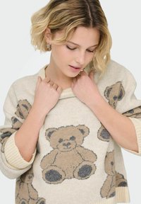 Beżowy sweter z dzianiny z wzorem misia w brązowym i szarym kolorze, z ściągaczami przy mankietach i szerokim dekoltem. Miękka tekstura.