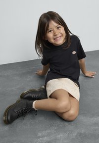 Camiseta de algodón negra con un pequeño logo, falda de algodón beige y botas de cuero negras con cordones y costuras amarillas sobre un suelo gris.
