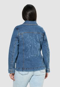 Džínová bunda ve středně modré barvě s přiléhavým střihem. Obsahuje grafiku na zadní straně, knoflíkové manžety a viditelné detaily šití.