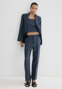 Persona joven con el cabello peinado hacia atrás, vestida con un traje de pantalón azul marino, top corto y sandalias de tacón negro, de pie contra un fondo blanco.