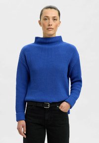 Blauer Strickpullover mit hohem Kragen, gerippter Textur und langen Ärmeln. Kombiniert mit schwarzen Jeans und einem schwarzen Gürtel.