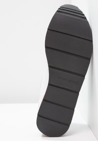 Semelle en caoutchouc noire avec une adhérence texturée, présentant des rainures horizontales ; tige de chaussure blanche avec une légère courbure et un accent rouge le long du bord.