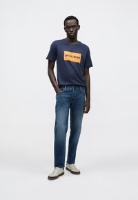 Μπλε ναυτικού χρώματος μπλουζάκι με πορτοκαλί τύπωμα λογοτύπου, συνδυασμένο με τζιν μεσαίου ξεβγάλματος και λευκά αθλητικά παπούτσια. Απλός σχεδιασμός, casual εφαρμογή.