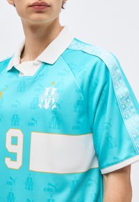 Isik, kes kannab helesinist spordisärki valge krae, numbriga 9, meeskonna logoga ja õlal dekoratiivse valge triibuga.