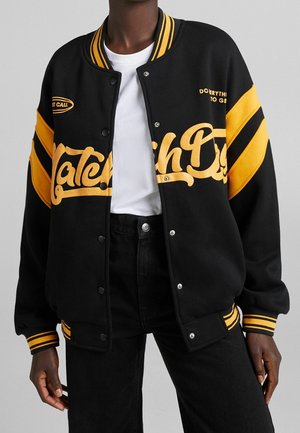 Zwarte varsity jacket met gele gestreepte mouwen, geborduurde letters op de voorkant en drukknopen. Draagt over een wit T-shirt.