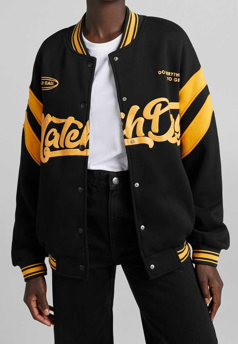 Svart varsityjacka med gula randiga ärmar, broderad text på framsidan och tryckknap stängning. Bärs över en vit t-shirt.