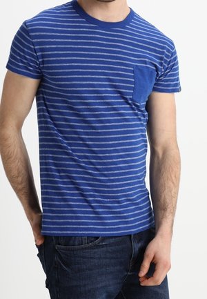 Mężczyzna ubrany w niebiesko-szarą pasiastą koszulkę z krótkim rękawem i kieszonką na piersi oraz ciemnoniebieskie dżinsy, stojący z jedną ręką w kieszeni.