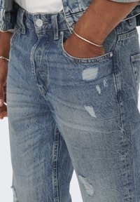 Vaaleansiniset farkut, joissa on kulutettu tyyli useilla revityillä kohdilla ja klassinen viiden taskun malli. Hopeanvärinen napin sulkeminen.