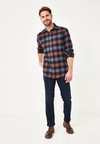 Mann in einem hochgekrempelten, karierten Hemd in Marineblau, Braun und Grau, dunklen Jeans und braunen Schnürboots, der vor einem schlichten weißen Hintergrund steht.