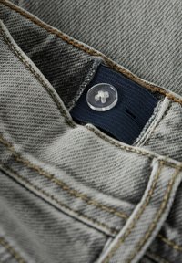 Pilkas džinsas su matomu mygtuku ir tamsiai mėlynu audinio etikete. Lengvai kontrastingos rudų siūlų siuvinėjimas ir tekstūruotas paviršius. Artimas vaizdas.