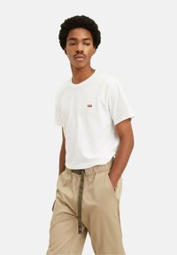 Ung mand med krøllet hår iført en hvid Levi's t-shirt og beige bukser med et synligt grønt bælte, hænderne i lommerne, ser til siden.