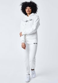 Felpa bianca con tasca frontale e cordoncino, abbinata a pantaloni sportivi bianchi coordinati. Accenti di logo neri su entrambi i capi. Sneakers bianche incluse.