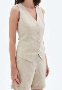 Beige linneväst med V-ringning, fem knappar och sidofickor, kombinerad med matchande skräddarsydda shorts. Mjuk textur, strukturerad design.
