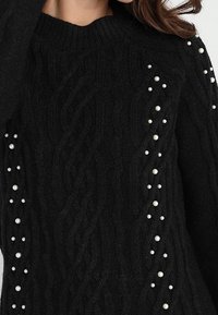 Czarny sweter z dzianiny z warkoczowymi wzorami i rozproszonymi białymi perłowymi ozdobami wzdłuż rękawów i przednich paneli.