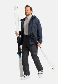 Skisetup: man in een marineblauwe weerbestendige jas, grijze skibroek, witte skischoenen, die witte ski's en trekkingstokken vasthoudt tegen een effen achtergrond.