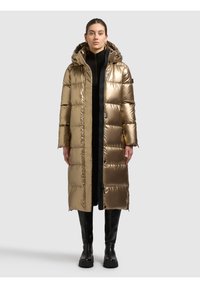 Metallic gouden donzen jas met capuchon, voorzien van een lang silhouet, doorgestikt ontwerp en een ritssluiting aan de voorkant. Draagt over een zwart outfit met hoge laarzen.