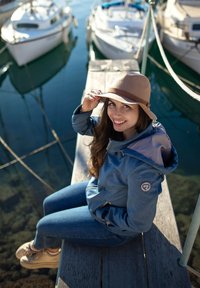 Denimjacke in hellblau mit braunen Akzenten und Logodetail. Die Person trägt einen beigen Hut und sitzt auf einem Holzsteg in der Nähe von Booten.