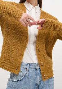 Senapsgul kofta i mjukt, texturerat stickat material med ribbade ärmslut och fem knappar. Bärs över en vit skjorta med krage och ljusblå jeans.