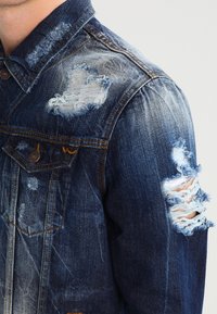 Denimjacka med slitna lappar på axeln och överarmen, med synliga fransade kanter och sömdetaljer.