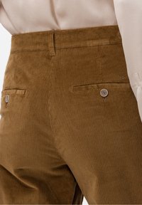 Bruna kordflodden byxor med vertikala ribbor, en platt midja och två bakfickor med knappstängning. Slät textur med en subtil lyster.