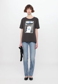 Grå bomulls t-skjorte med trykket grafisk design av et ansikt, kombinert med lyseblå jeans og svarte sko.