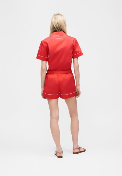 Ξανθιά γυναίκα που φοράει κόκκινο κοντομάνικο πουκάμισο και ταιριαστά σορτς με λευκή επένδυση, στέκεται ξυπόλυτη σε λευκό φόντο, γυρισμένη προς τα πίσω.