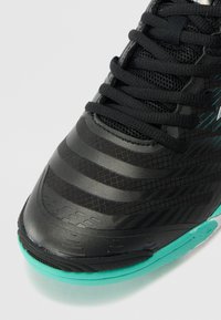 Black athletic shoe with mesh and textured accents, featuring turquoise sole and laces, contoured design, and striped details on the upper.