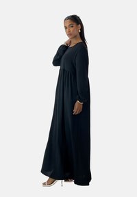 Vestido negro de manga larga con cintura fruncida, silueta fluida, hecho de tela ligera, combinado con sandalias de tacón plateadas.