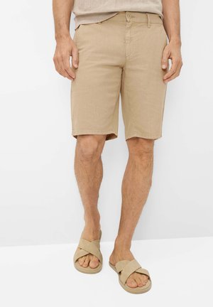 Man draagt beige knielange shorts en beige sandalen met gekruiste bandjes, staand tegen een effen witte achtergrond.