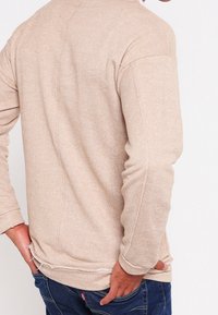 Beige svetr s dlouhým rukávem, texturovaná tkanina, žebrované manžety a roztrhaný lem. Zobrazeno ze zadní strany, zvýrazňující uvolněný střih.