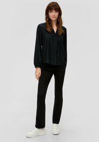Schwarze Bluse mit langen Ärmeln und V-Ausschnitt, aus leichtem Stoff, etwas lockere Passform und gerüschte Manschetten. Kombiniert mit schwarzen Slim-Fit-Hosen und weißen Sneakern.