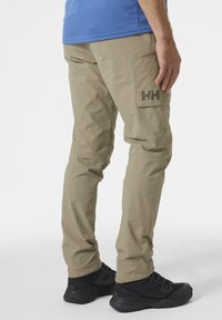 Beige cargobroek met een zijzak, featuring een donker logo. Draagt met zwarte wandelschoenen. Glad materiaal en een ontspannen pasvorm.