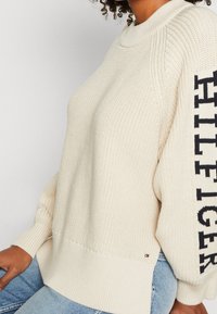 Femme portant un pull en tricot crème avec "HILFIGER" en lettres noires sur la manche et un jean bleu clair, assise.
