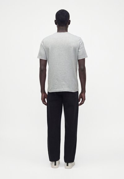 Homme debout, de dos, portant un t-shirt gris à manches courtes, un pantalon noir et des baskets blanches sur un fond clair uni.
