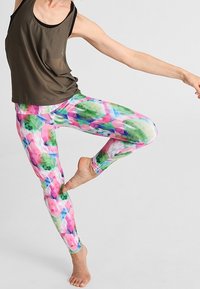 Kolorowe legginsy sportowe w różowe, zielone i białe wzory kwiatowe, zestawione z luźną brązową bluzką bez rękawów wykonaną z oddychającego materiału.