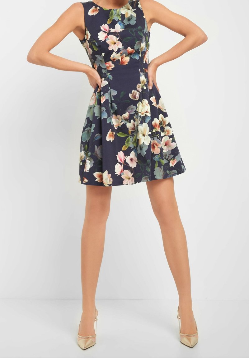 Robe évasée fleurie bleu marine avec de grands motifs floraux multicolores. Sans manches, longueur au genou, assortie à des talons métallisés.