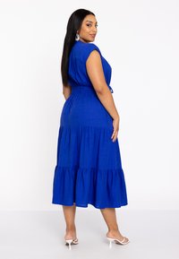 Blaues, gestuftes Maxikleid aus gewebtem Stoff, mit kurzen Ärmeln, einer taillierten Taille und einem fließenden Saum. Mit Taschen akzentuiert.