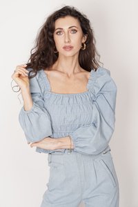 Jasnoniebieska denimowa bluzka z bufiasnymi rękawami i kwadratowym dekoltem z marszczonymi detalami. Modelka ma na sobie spodnie dżinsowe z wysokim stanem.