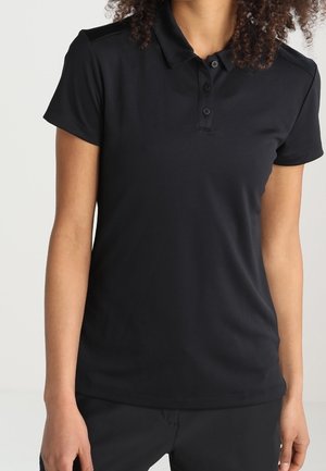 Persoon met een zwart poloshirt met korte mouwen en een zwarte broek, staand tegen een lichte achtergrond, zichtbaar van schouders tot heupen.